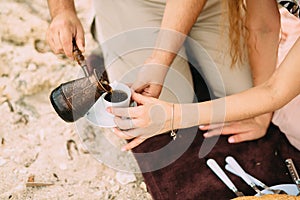 Hands man pours turkish coffee from a metal coffee pot into a white cup and saucer, women`s hands help