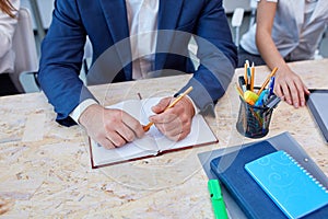 Hands of a man in a suit is sitting at desk and holding an pencil next to a notepad.