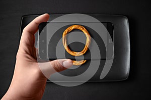 Hands of a man takes photos of food on the table with the phone. Bagel on a black plate. Dry bread ring, cracker on a black