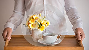 The hands of a man in a white shirt are holding a tray with a cup of coffee / tea and a bouquet of daisies against his chest