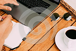 Hands of a man working at a laptop top view of a workspace, on a wooden table with a cup of coffee and a notebook for recording