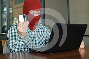 Hands of masked hacker wearing a balaclava holding credit card between stealing data from laptop. Internet crime concept.