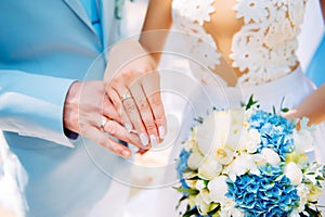 Hands of newlyweds with beautiful gold rings, close-up. White bridesmaid dress, bouquet, stylish manicure. Perfect wedding