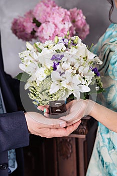 The hands of the newlyweds hold a box-case with gold wedding rings and a bouquet of flowers. A bouquet of white cream roses and