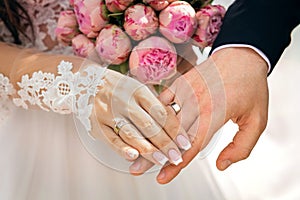 Hands of the newlyweds with rings on the fingers, next to a bouquet with pink peonies, the bride and groom hold hands