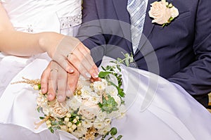 The groom`s hands with wedding rings lie on top of each other, on the bride`s bouquet of roses