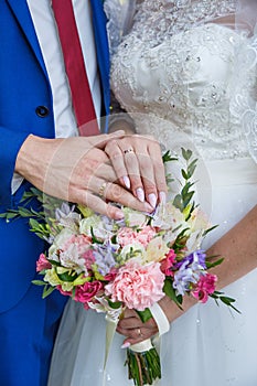 Hands of newlyweds with wedding rings and a bouquet of flowers. The groom in a blue suit and red tie. The concept of