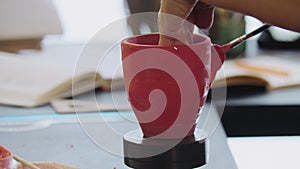 Hands of an old woman painting clay mug in red color