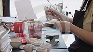 Hands of an old woman painting a little clay cup with dusty pink color