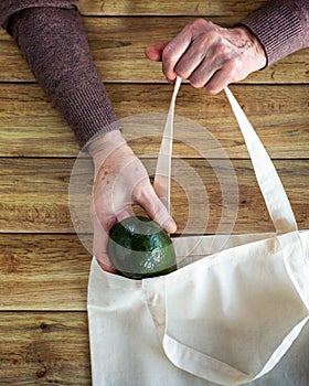 Hands of an old woman put fresh organic avocado in Eco Shopping Bag on wooden background, Flat Lay