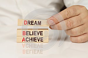 In the hands of a person and on a reflective surface are wooden blocks with the inscription - Dream Believe Achieve
