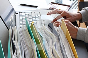 Hands of person typing on laptop computer with binders filled with papers in foreground. Selective focus