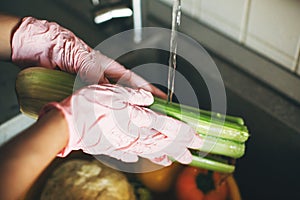 Hands in pink gloves washing celery in water stream in sink during virus epidemic. Woman  cleaning fresh vegetables, preparing for