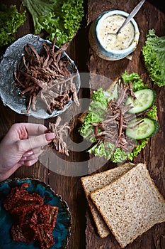 Hands preparing sandwich with meat, dark bread, fresh green salad, dry tomatoes and fresh cucumbers on rustic wooden table surface