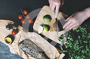 Hands in the process of cooking fish, pepper, parsley, tomato, lime on a cutting board on a black wooden background top view horiz