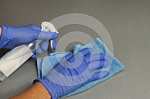 Hands in protective gloves cleaning and sanitizing gray desk table surface at office or home with antibacterial spray and cloth.