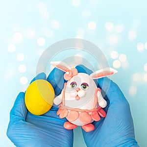 Hands in protective medical gloves holding a painted yellow Easter egg and a homemade Easter bunny