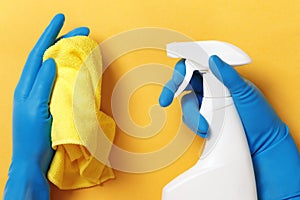 Hands in protective rubber gloves holding a spray bottle with detergent and a cloth on a yellow background. Hygiene prevention.