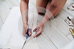 Hands with scissors with blue handles, cutting paper pattern with light beige fabric. Close-up picture of cutting process. Sewing