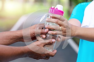 Hands of senior man holding bottle of fresh water for his wife while workout in the park