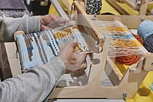 Hands of senior man weaving small rug with pattern on manual table loom.