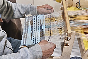 Hands of senior man weaving small rug with pattern on manual table loom.