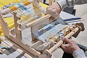 Hands of senior man weaving small rug with pattern on manual table loom, at masterclass on weaving.