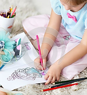 Hands of small girl in home clothing sitting on soft carpet and coloring doll drawing with colorful pencils at home