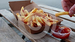 Hands taking french fries from box and dipping them in ketchup on wooden table - closeup, natural day light, otside