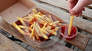 Hands taking french fries from box and dipping them in ketchup on wooden table - closeup, natural day light, otside