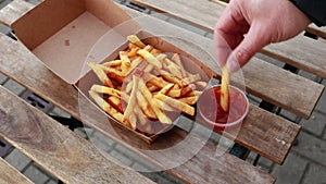 Hands taking french fries from box and dipping them in ketchup on wooden table - closeup, natural day light, otside
