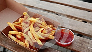 Hands taking french fries from box and dipping them in ketchup on wooden table - closeup, natural day light, otside