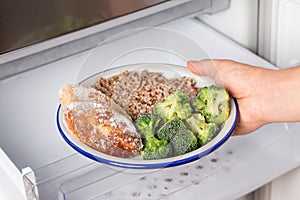 Hands are taking a plate of frozen cutlets and vegetables out of the freezer of the fridge. Concept of storing ready made food,