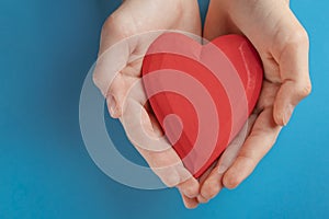 Hands of a teenager child holding a red wooden heart in their hands. Blue background