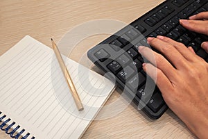 Hands typing keyboard with a blank notepad and pencil on wooden