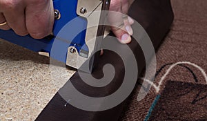 Hands upholstering the paricle board using blue stapler and brown leather.