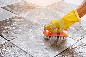 Hands wearing yellow rubber gloves are using a plastic floor scrubber to scrub the tile floor.