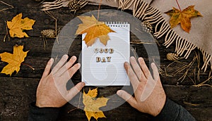 Hands and White blank notebook with wooden letters phrase Good day on a dark background with a scarf, plaid and cup, autumn yellow