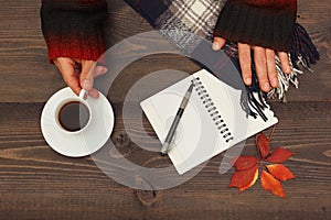 Hands of woman with a cup of coffee and scarf at wooden table with notebook and pen and autumn leaves