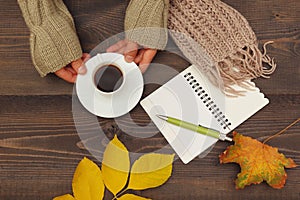 Hands of woman with a cup of espresso and scarf at wooden table with notebook and pen and autumn leaves