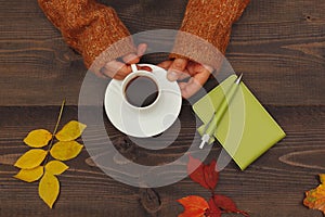 Hands of woman with a cup of espresso at a wooden table with notebook and pen and autumn leaves