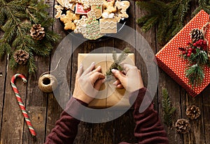 Hands of a woman decorating a Christmas gift with conifer branch on rustic wooden table full with cookies in festive winter