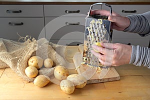 Hands of a woman are grating potatoes with a metal grater on a wooden kitchen table