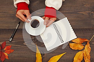 Hands of woman hold a cup of coffee at a wooden table with notebook and pen and autumn leaves