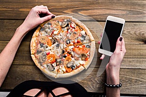 Hands of a woman ordering pizza with a device over a wooden workspace table. All screen graphics are made up.