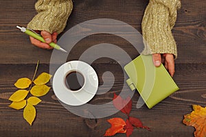 Hands of woman with a pen and notebook at a wooden table with cup of coffee and autumn leaves