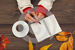 Hands of woman with a pen and notebook at a wooden table with cup of espresso and autumn leaves