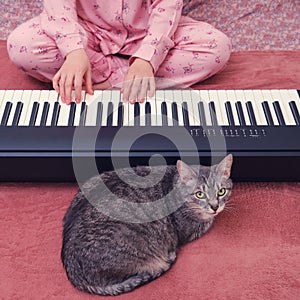 The hands of a woman in pink pajamas on the keys of an electronic piano next to a cat