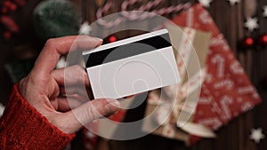 Hands of a woman in a red sweater with a credit card on a background of gift boxes, selective focus, close-up. Christmas and New