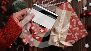 Hands of a woman in a red sweater with a credit card on a background of gift boxes, selective focus, close-up. Christmas and New